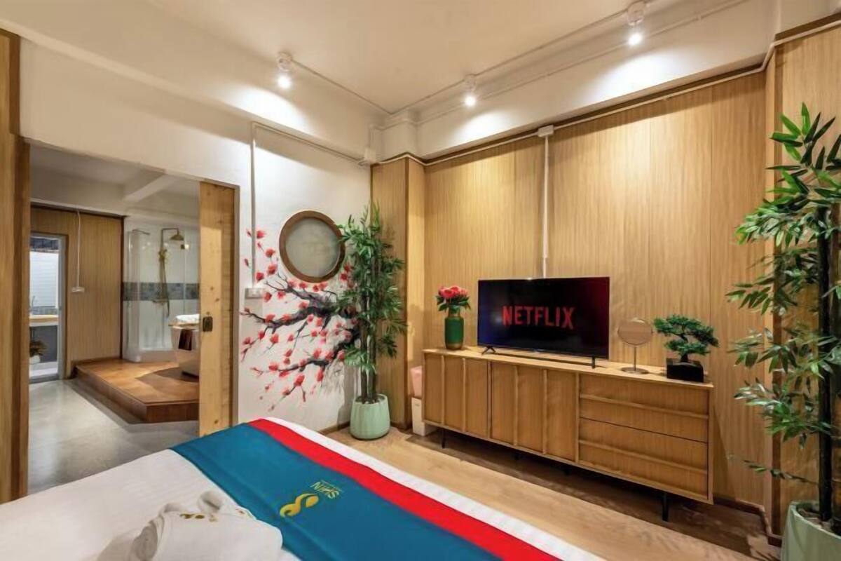 A modern hotel room with a bed, a wooden TV stand, a wall-mounted TV, a mirror, and a potted plant.