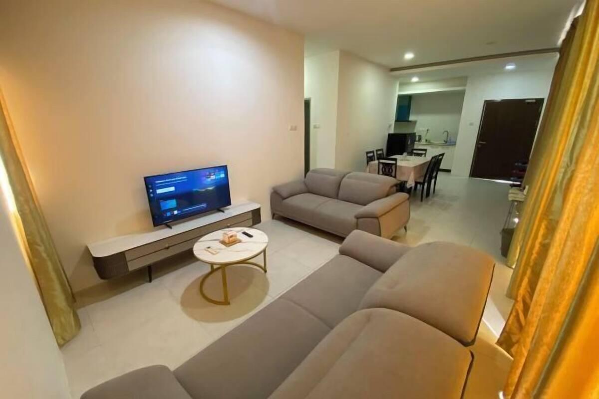 A modern living room with a flat-screen TV, a sofa, a coffee table, and a dining area in the background.