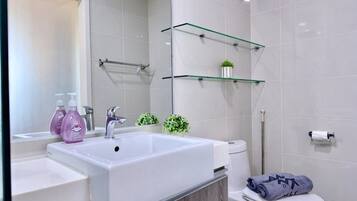 A bathroom with a white sink, mirror, and toilet. There are two glass shelves above the sink, one holding a plant and the other a towel.