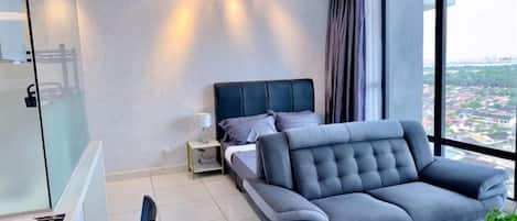 Une chambre moderne avec un grand lit, un canapé gris, une table de chevet et une vue sur la ville grâce à de grandes fenêtres.