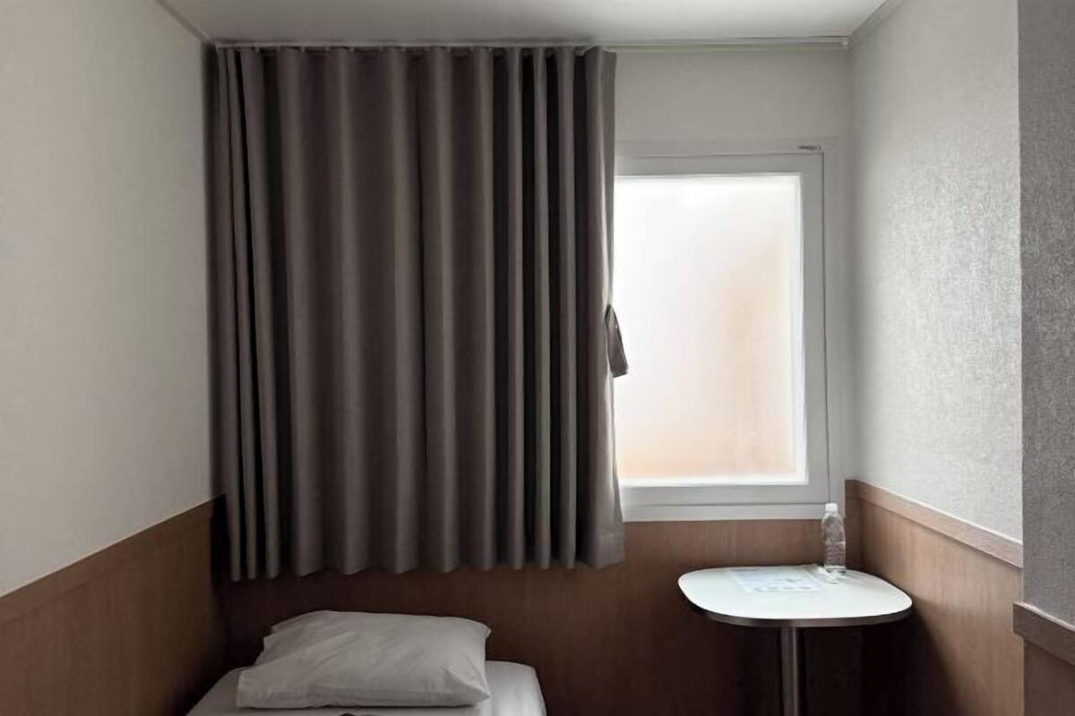 A hotel room with a bed, a small table, a window with curtains, and a bottle of water.