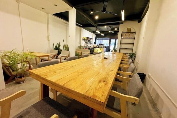 A long wooden table with chairs, a potted plant, and a shelf with books in a room with white walls and a black ceiling. - Truman Hostel (Luang Prabang)
