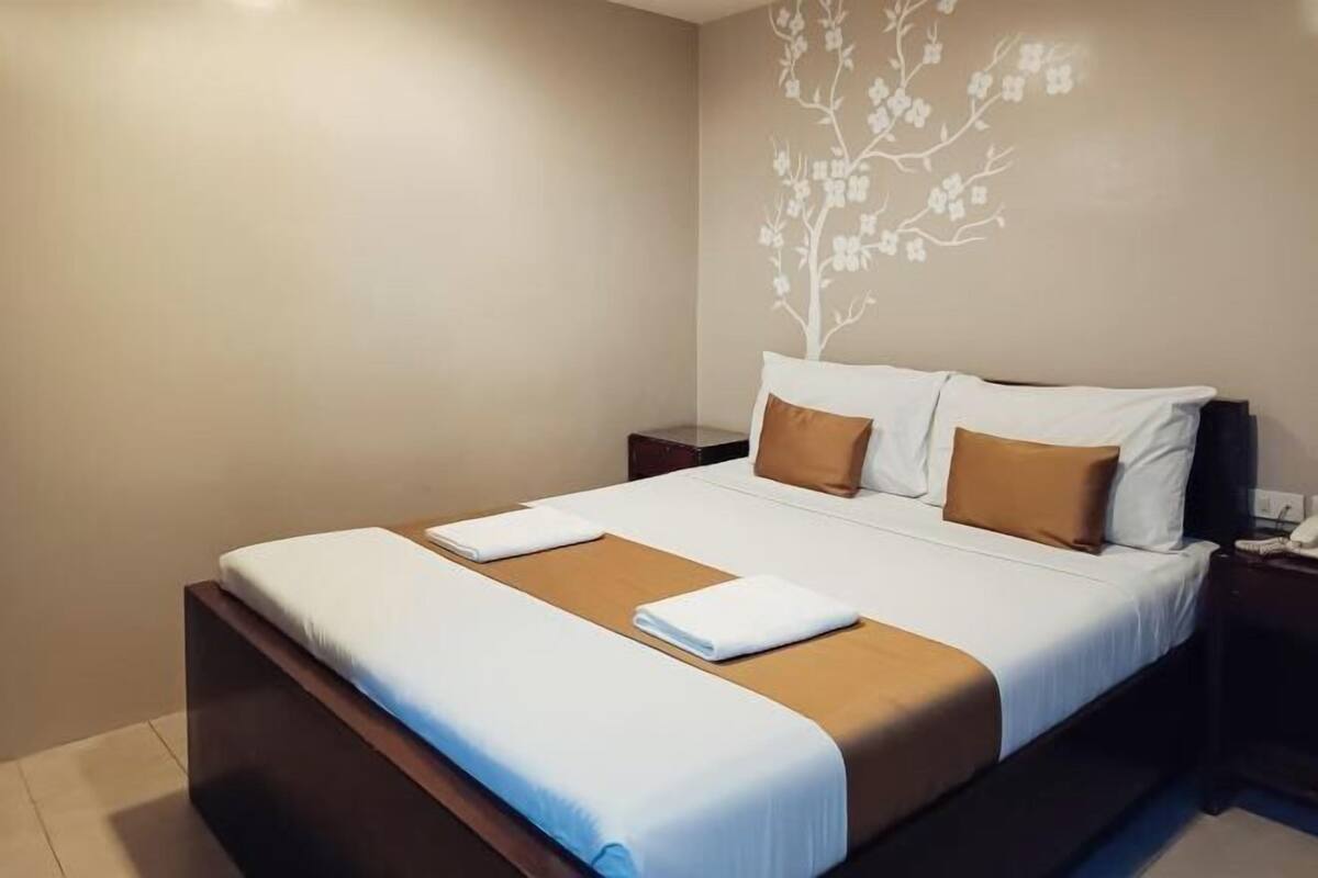 A neatly made bed with white and brown bedding, a tree decal on the wall, and a small bedside table with a lamp.