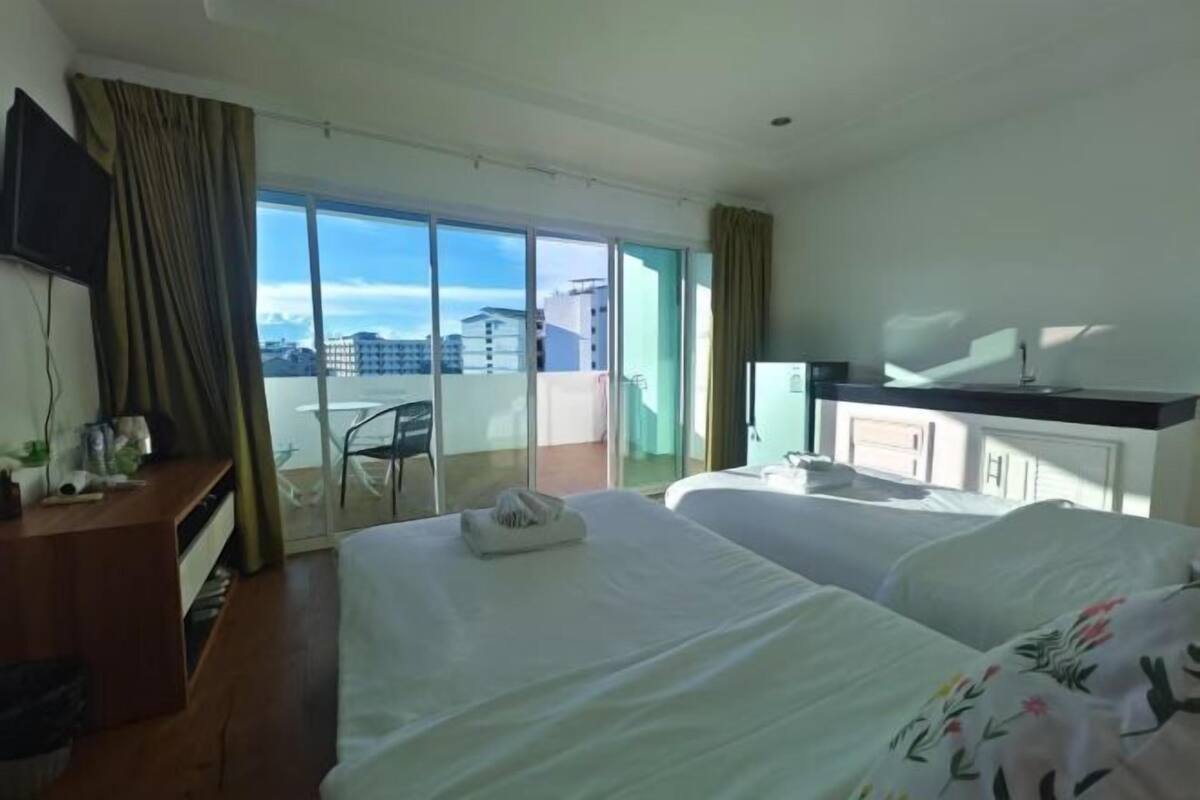 A hotel room with two beds, a TV, a desk, and a balcony with a view of buildings.
