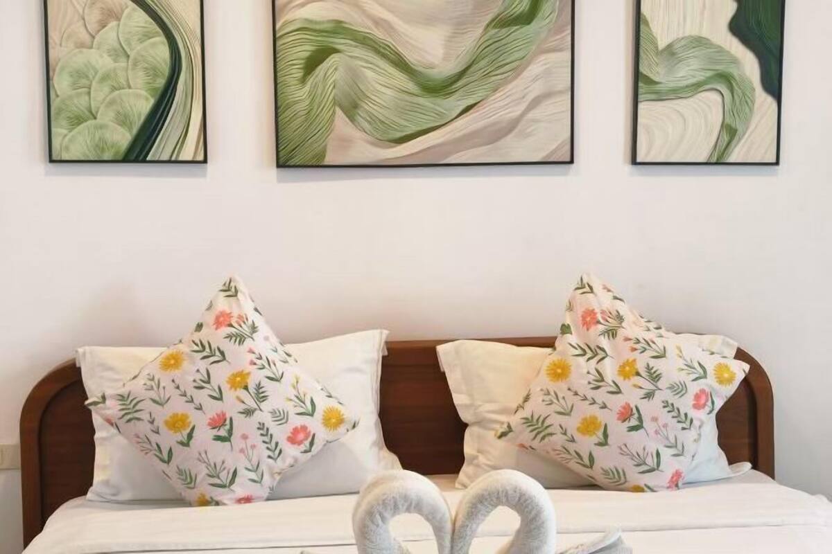 Un lit agrémenté d’oreillers à motifs floraux, une serviette en forme de cœur et une œuvre d’art abstraite accrochée au mur.