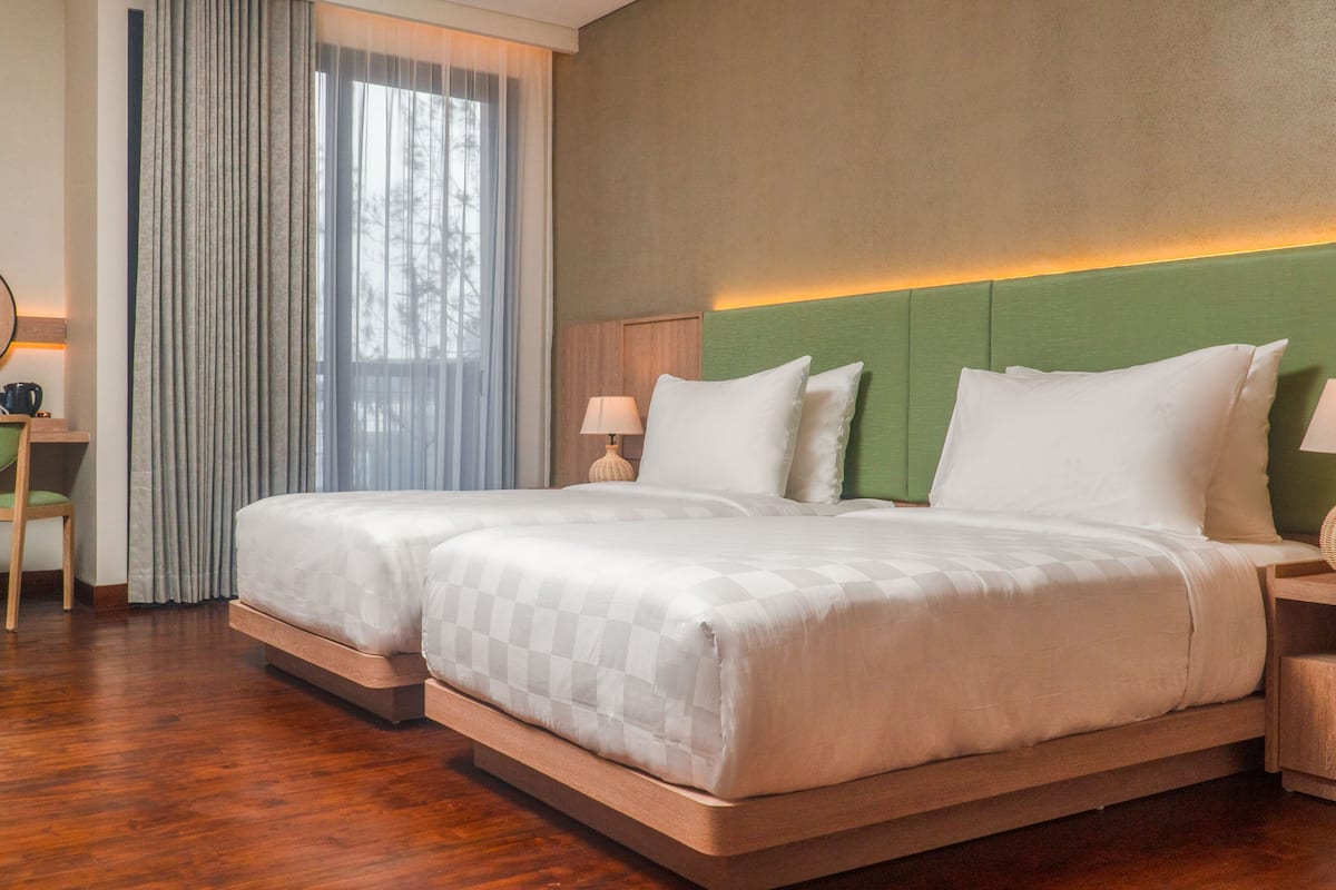 Una cama doble con cabecera verde, dos mesitas de noche con lámparas, un escritorio de madera con silla y un ventanal con cortinas.