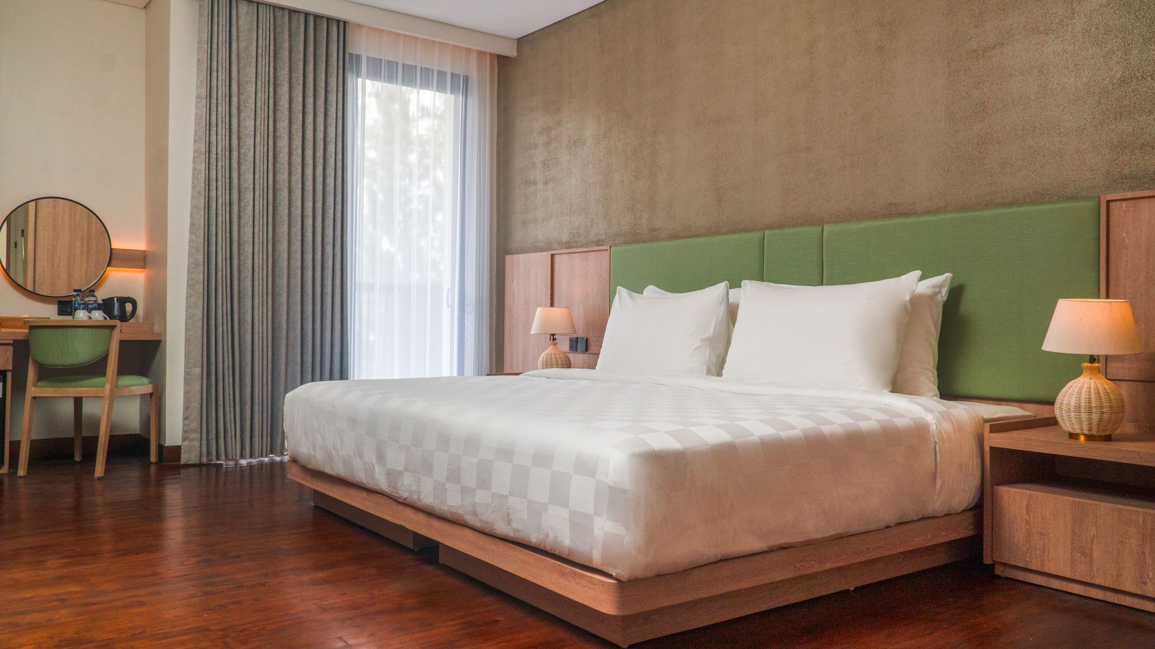 A hotel room with a large bed, a desk with a chair, a bedside table with a lamp, and a window with curtains.