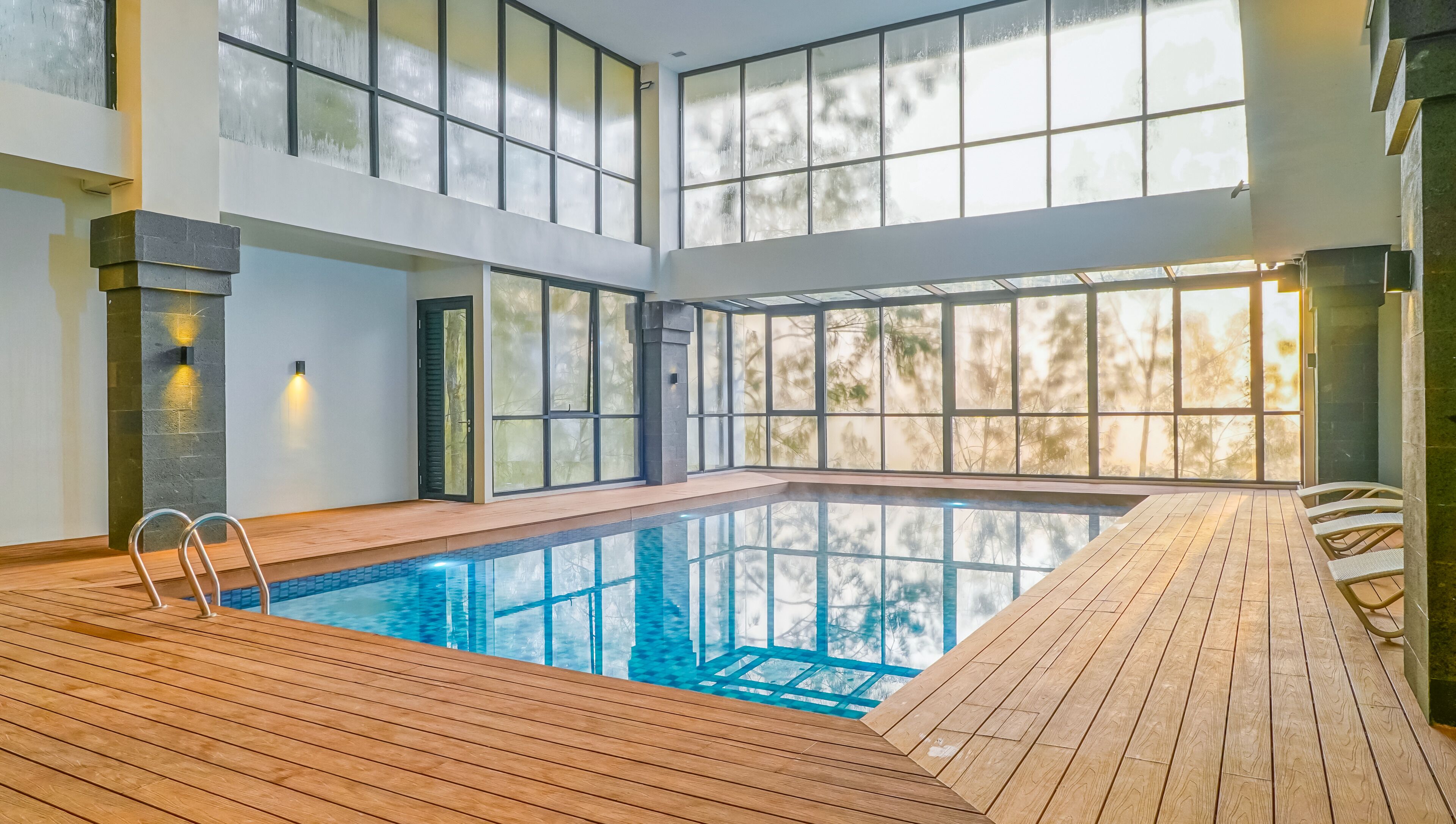 An indoor swimming pool with wooden decking, large windows, and a view of trees.