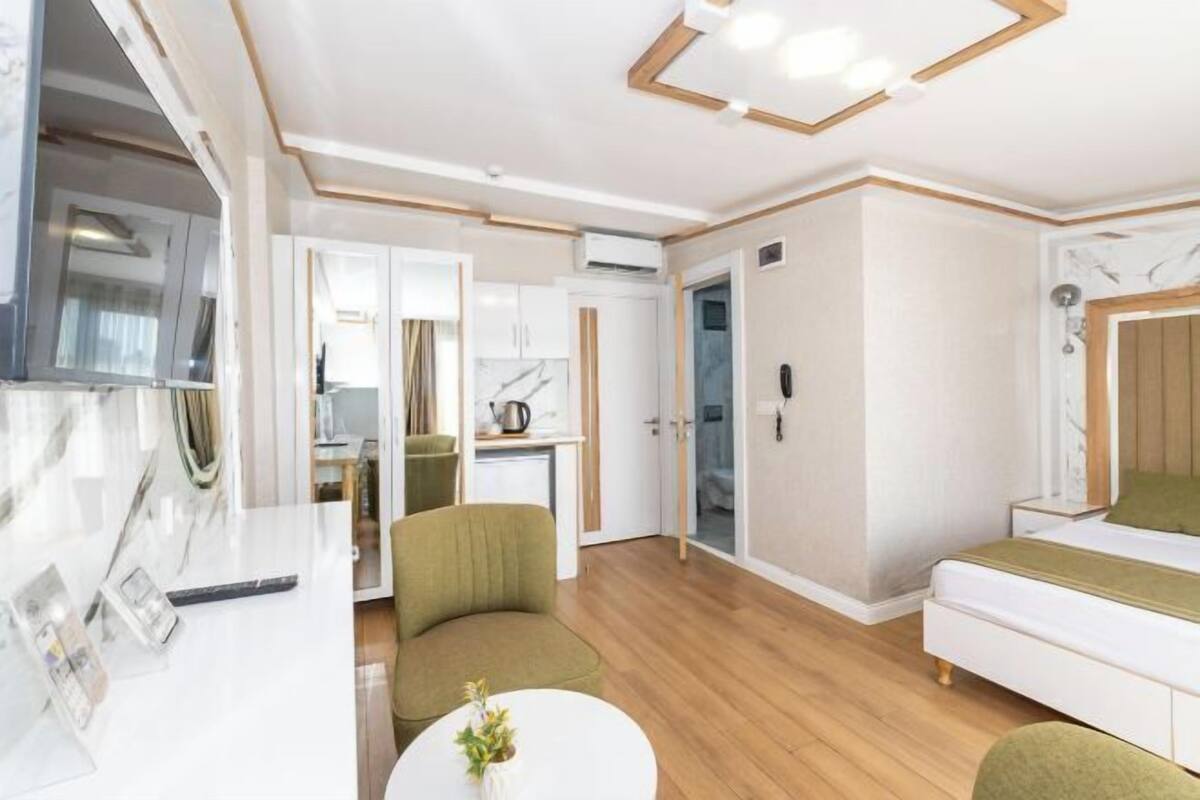 A modern hotel room with a wooden floor, a green armchair, a white sofa, a small round table with a vase, and a bed with a green pillow.
