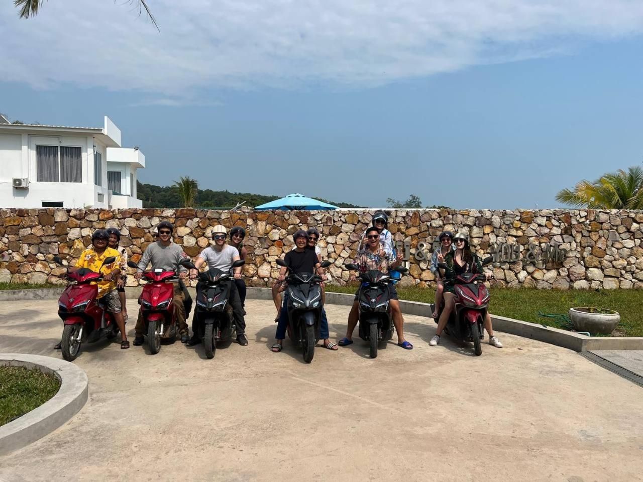 A group of people on motorcycles in a paved area with a stone wall and a building in the background.