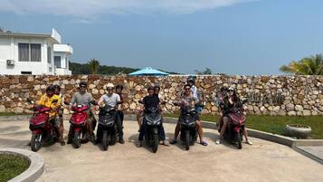 A group of people on motorcycles in a paved area with a stone wall and a building in the background.