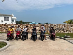 Un grupo de personas en motocicletas en una zona pavimentada, con un muro de piedra y un edificio al fondo.