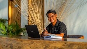 A person seated at a wooden table with a laptop and a notebook, in a room with a wooden wall and indoor plants. - You Me Resort (Koh Rong)