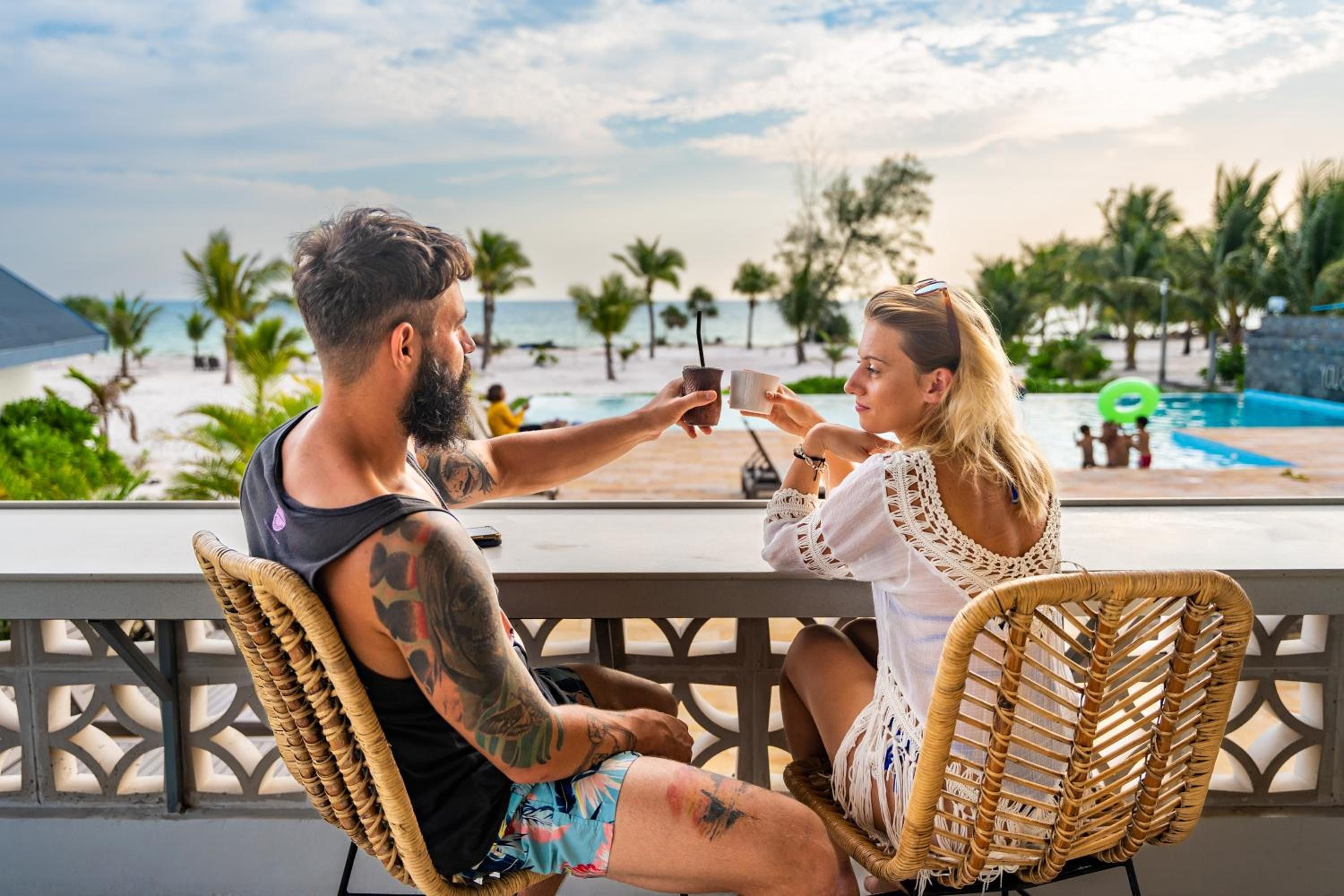Two people seated on wicker chairs facing a pool with a view of the beach and palm trees.