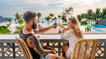 Two people seated on wicker chairs facing a pool with a view of the beach and palm trees.