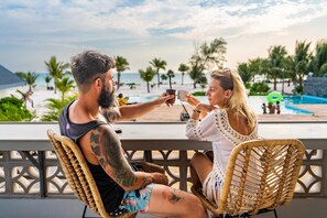 Dos personas sentadas en sillas de mimbre mirando una piscina con vista a la playa y palmeras.