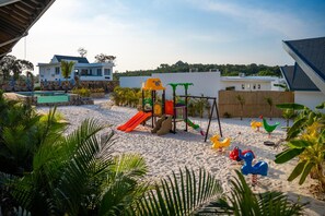 A sandy playground area with slides and swings, surrounded by palm trees and a pool. - You Me Resort (Koh Rong)