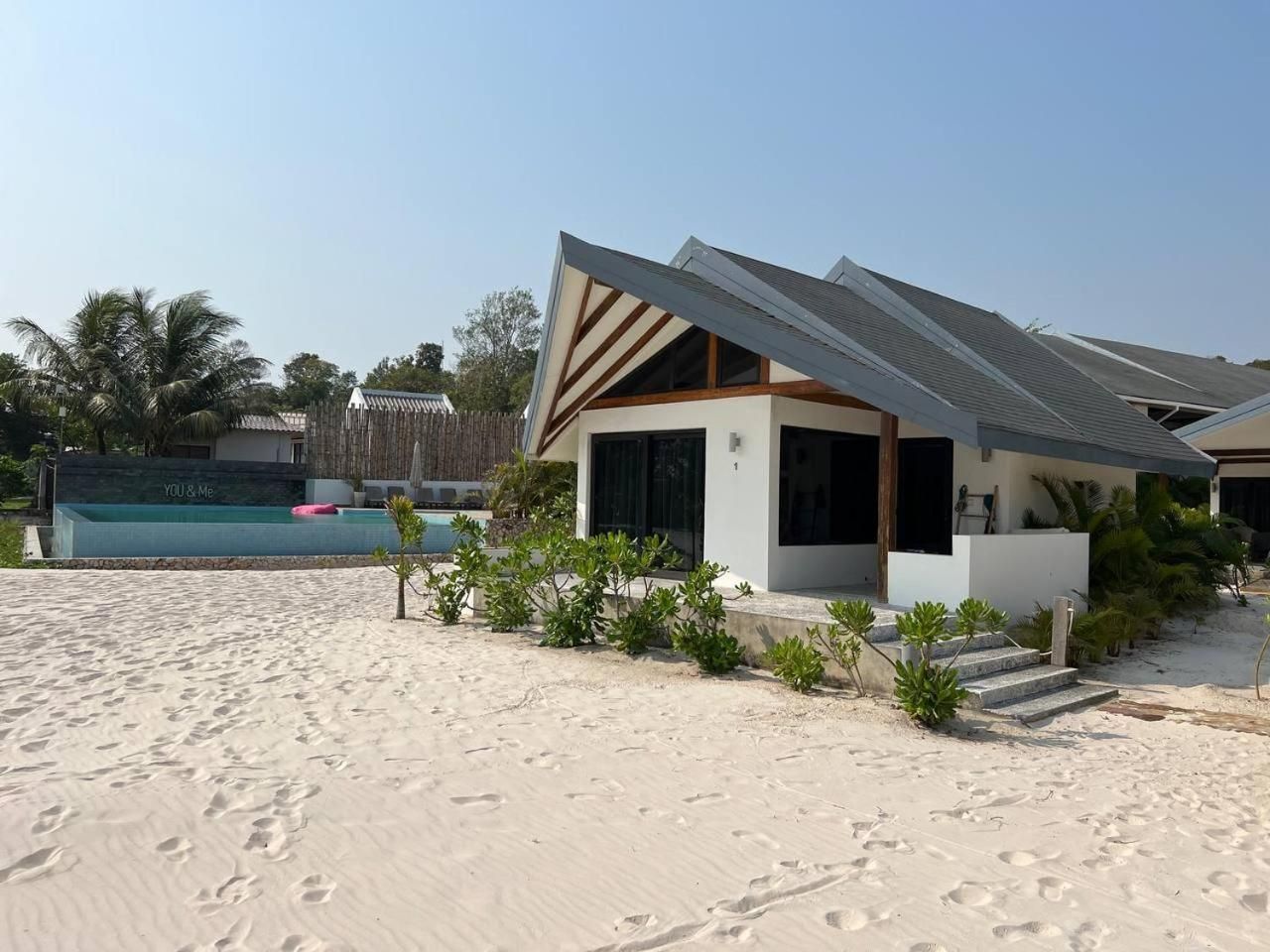 Een modern strandhuis met een zwembad, omringd door zand en groen.