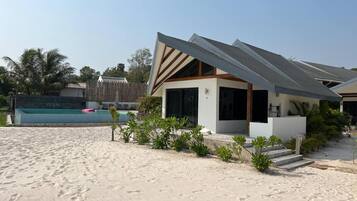 Een modern strandhuis met een zwembad, omringd door zand en groen.