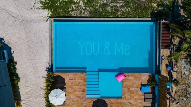 An aerial view of a swimming pool with 'YOU & Me' written on the water, surrounded by a wooden deck and umbrellas. - You Me Resort (Koh Rong)