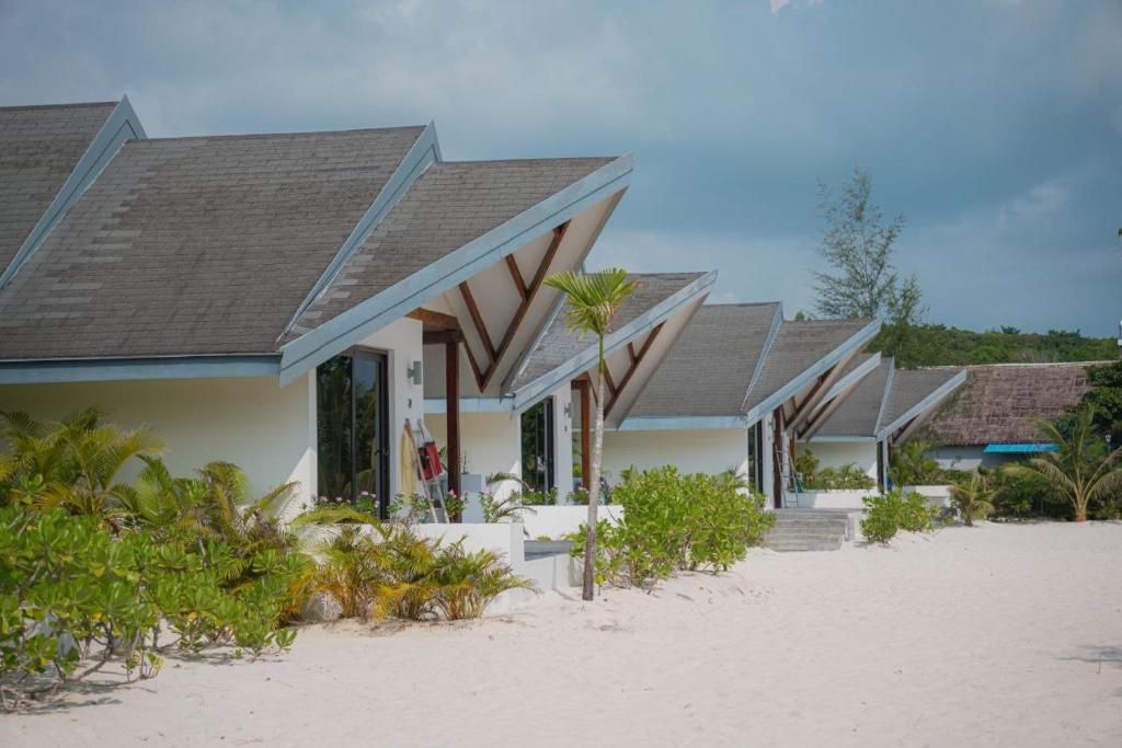 Ein Strandhotel mit mehreren Zimmern, das sich durch schräge Dächer und einen Sandstrand im Vordergrund auszeichnet.