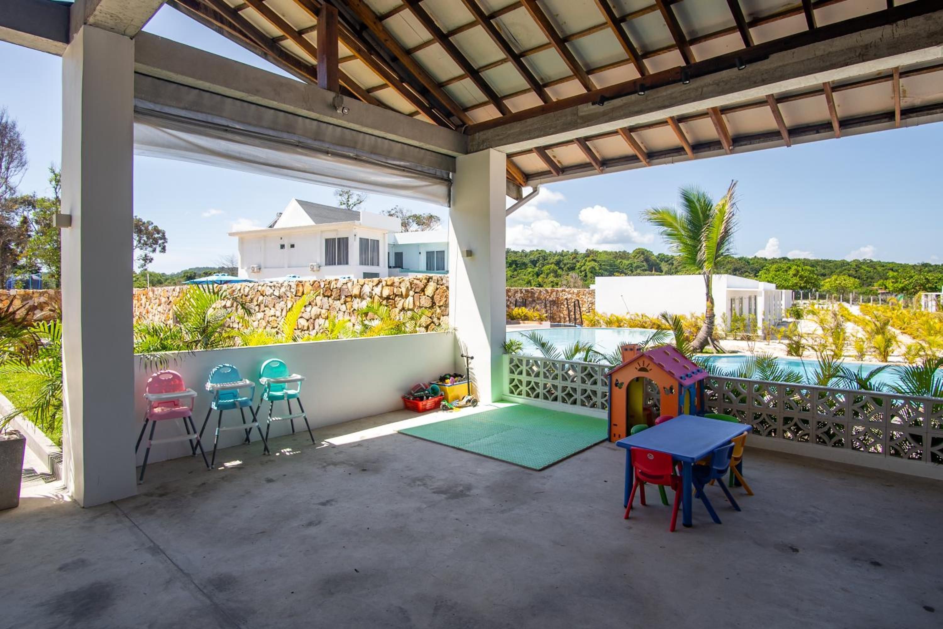 A covered outdoor area with a concrete floor, a small table with chairs, a children's playhouse, and a view of a white building and greenery.