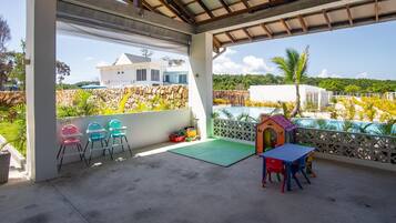A covered outdoor area with a concrete floor, a small table with chairs, a children's playhouse, and a view of a white building and greenery.
