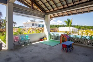 Un espacio al aire libre techado con piso de concreto, una mesa pequeña con sillas, una casita de juegos para niños, y vistas a un edificio blanco y vegetación.