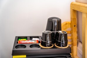 A coffee maker with two cups on a black tray, a packet of snacks, and a wooden chair in the background. - You Me Resort (Koh Rong)