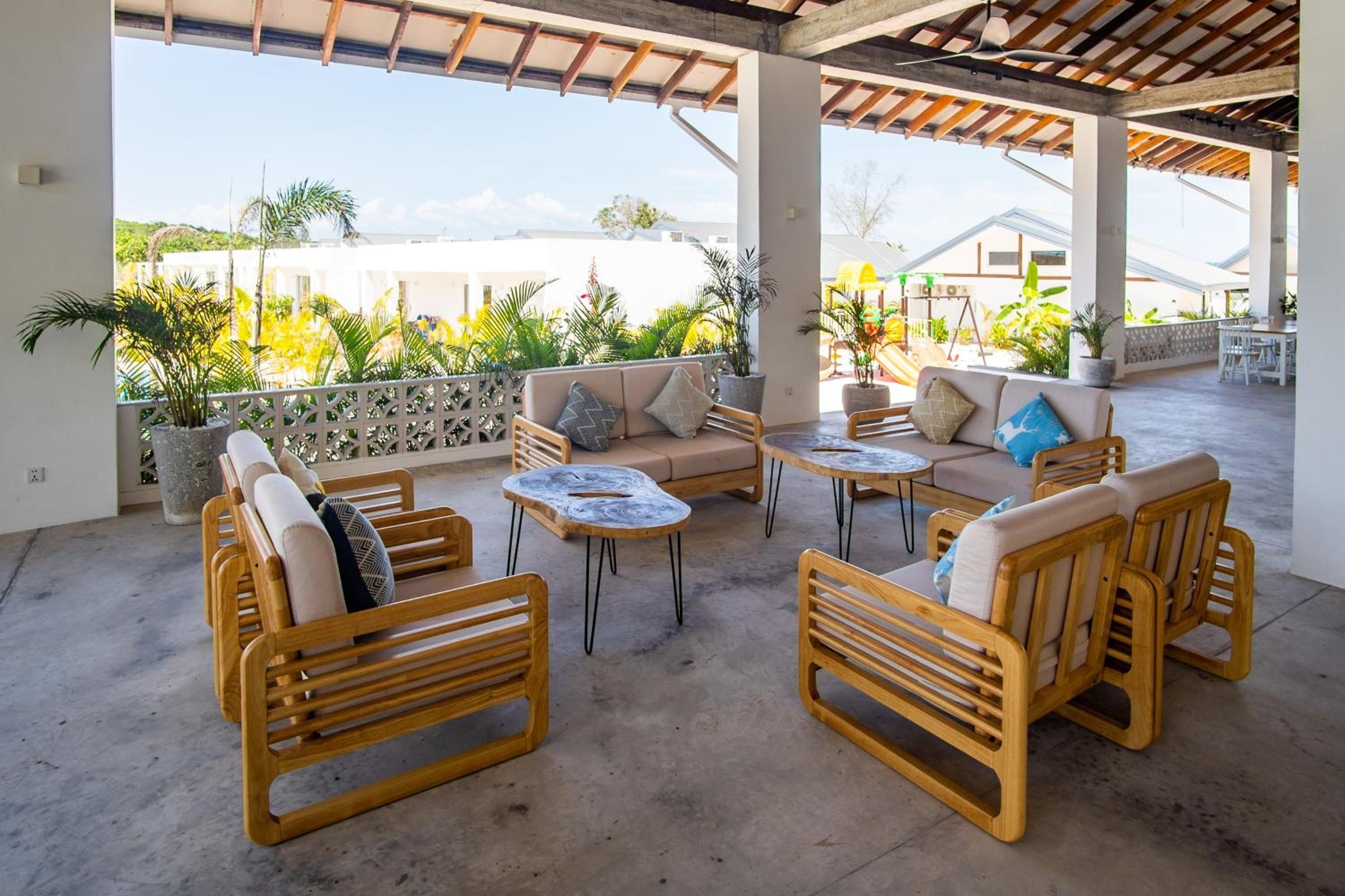 An outdoor seating area with wooden furniture, a concrete floor, and a view of greenery and buildings in the background.