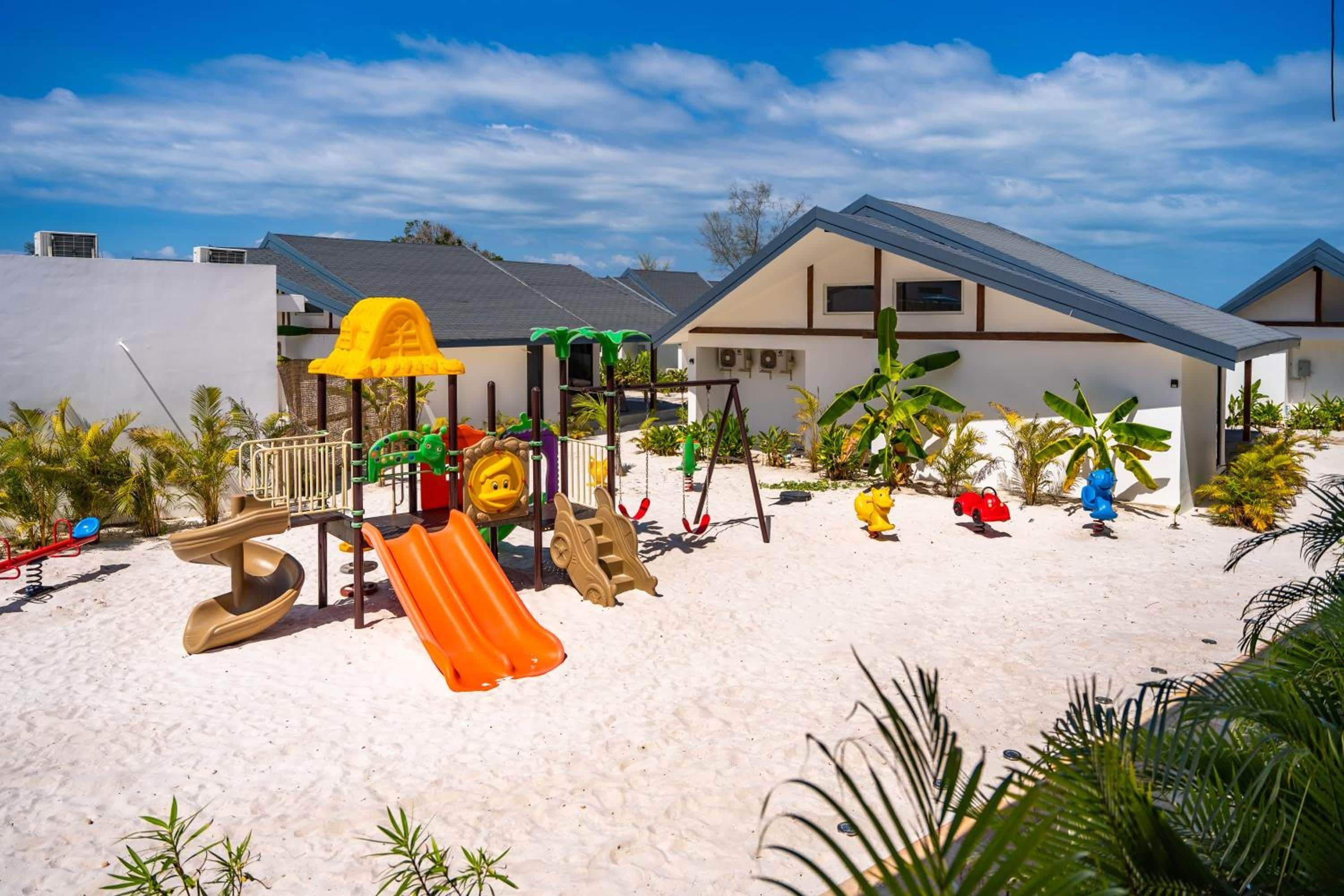 Un parque infantil con toboganes y columpios, rodeado de palmeras y un edificio con techo inclinado.