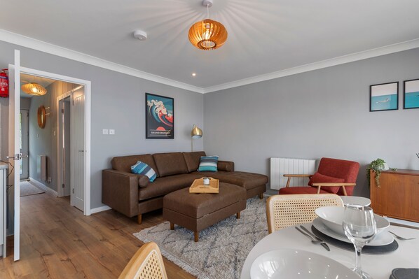 A modern living room with a brown sectional sofa, a dining table set for two, and a red armchair. - 33 Beach Court - Direct Beach Access Sea Views (Saundersfoot)