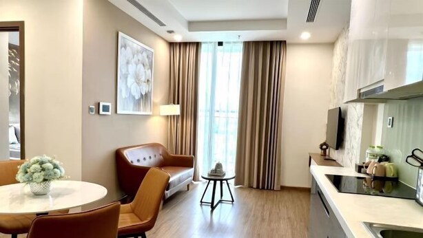 A modern hotel room with a dining area, a sofa, a small table with a lamp, and a kitchen area with a sink and countertop. - HKG - Vinhome GreenBay - Green apartment in Hanoi (Hanoi)