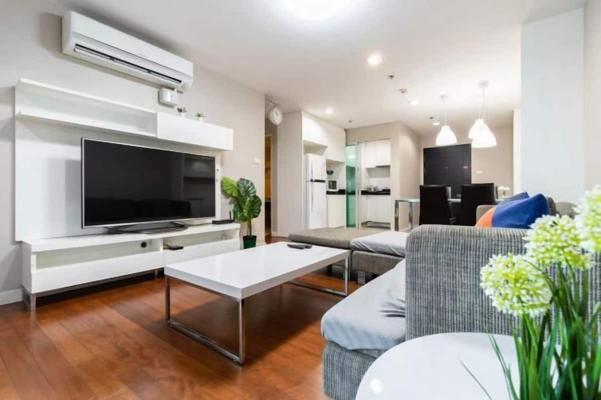 A modern living room with a flat-screen TV, a white coffee table, a grey sofa, and a dining area with pendant lights.