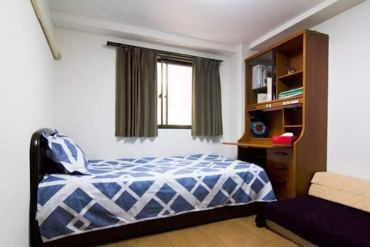 Un lit simple avec une couvre-lit à motifs géométriques, une armoire en bois avec des étagères, une fenêtre avec des rideaux et un petit bureau avec une chaise.