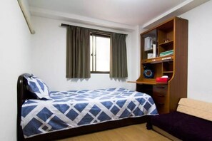 Un lit simple avec une couvre-lit à motifs géométriques, une armoire en bois avec des étagères, une fenêtre avec des rideaux et un petit bureau avec une chaise.
