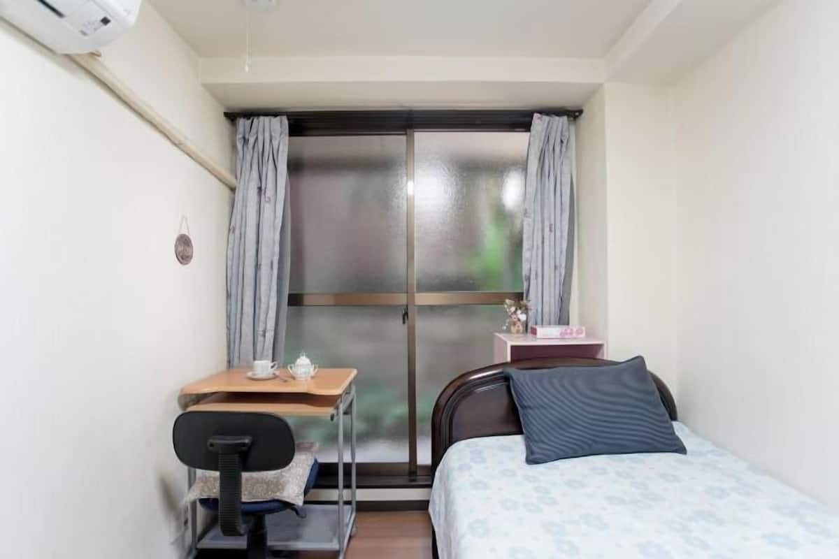 Une petite pièce, bien rangée, avec un lit, un bureau et une chaise, et une fenêtre avec des rideaux.