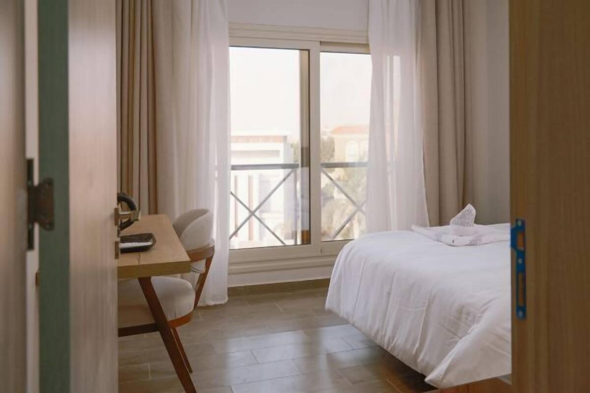 Une chambre à coucher avec un lit, un bureau et une chaise, une fenêtre avec des rideaux et une vue sur des immeubles.