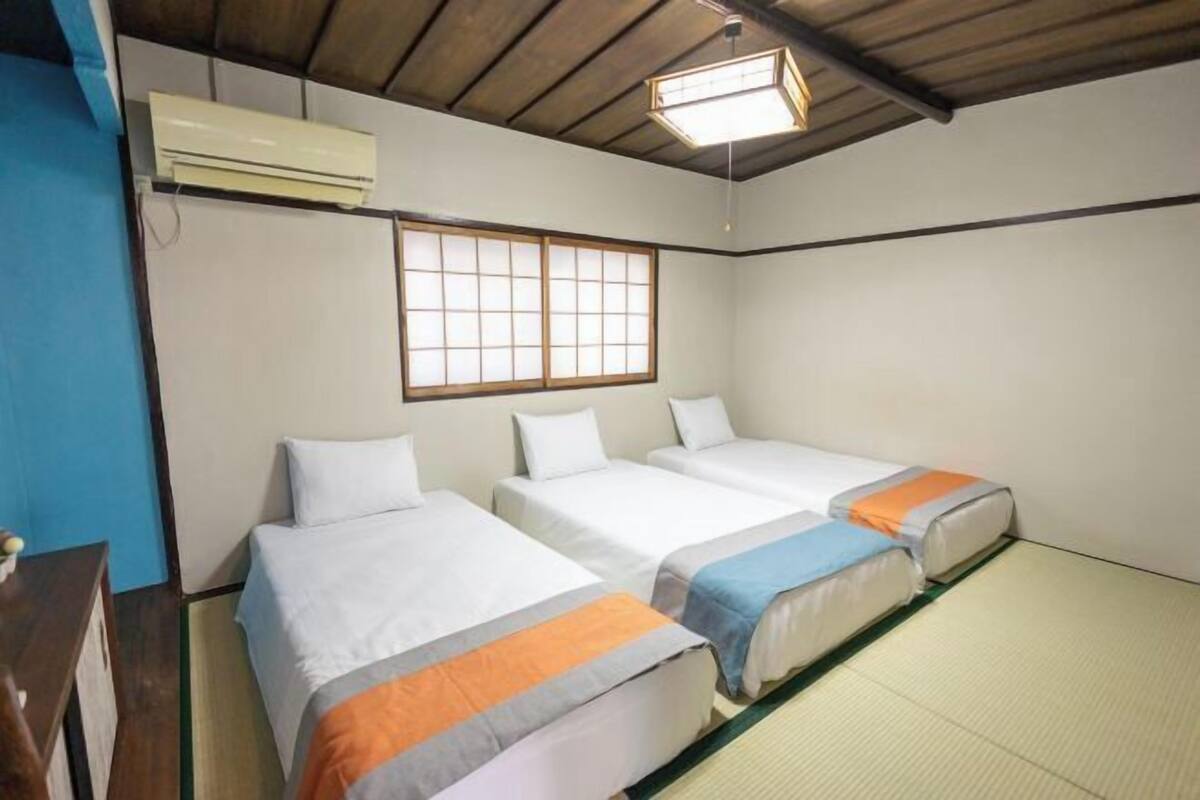 Una habitación con tres camas, una ventana de madera, un equipo de aire acondicionado y una lámpara en el techo.