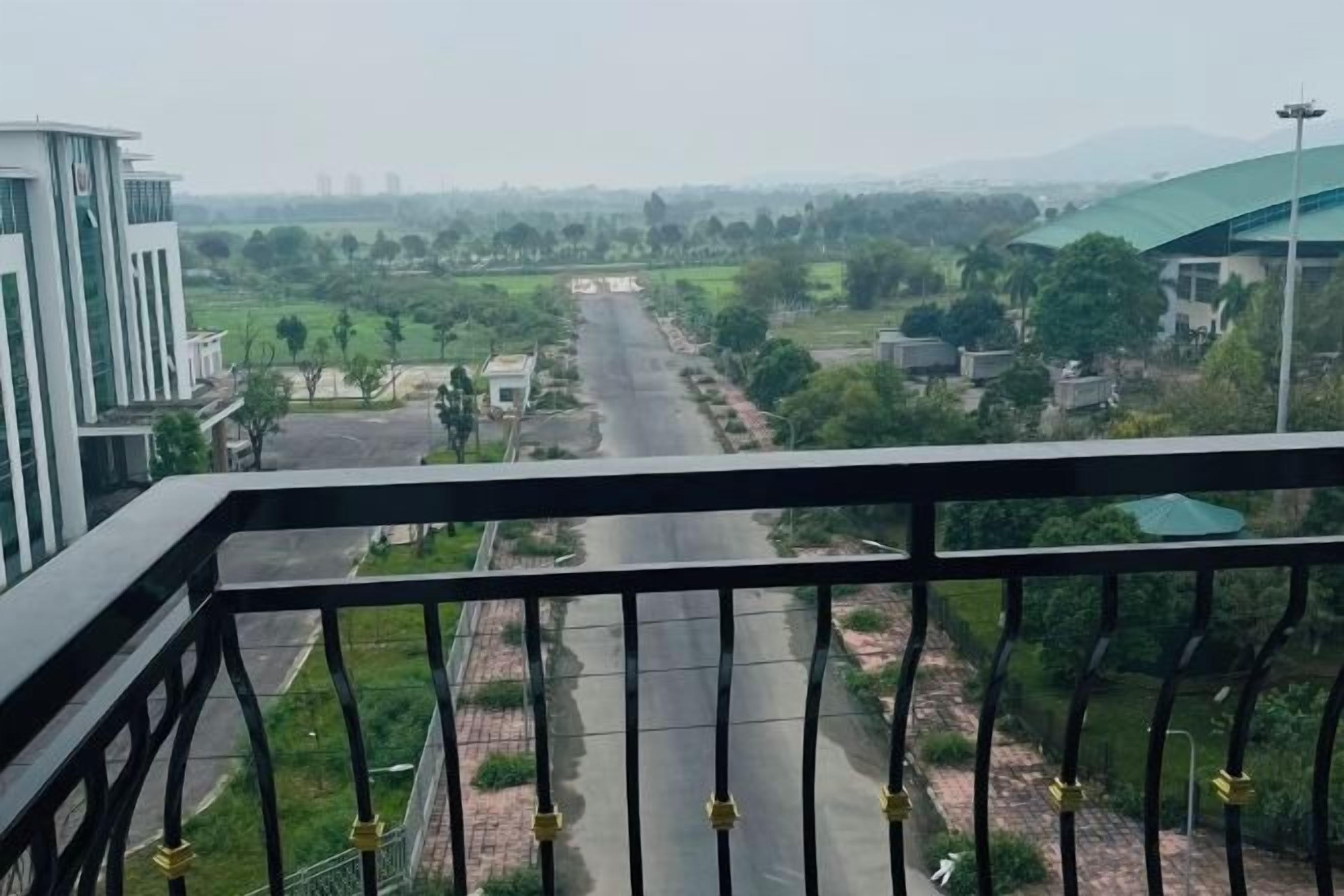 A view from a balcony overlooking a road with buildings and greenery in the distance.