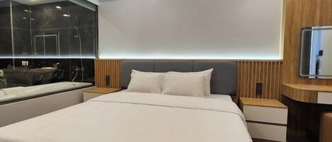 A modern hotel room with a large bed, a built-in TV, a vanity with a mirror, and a bathtub.