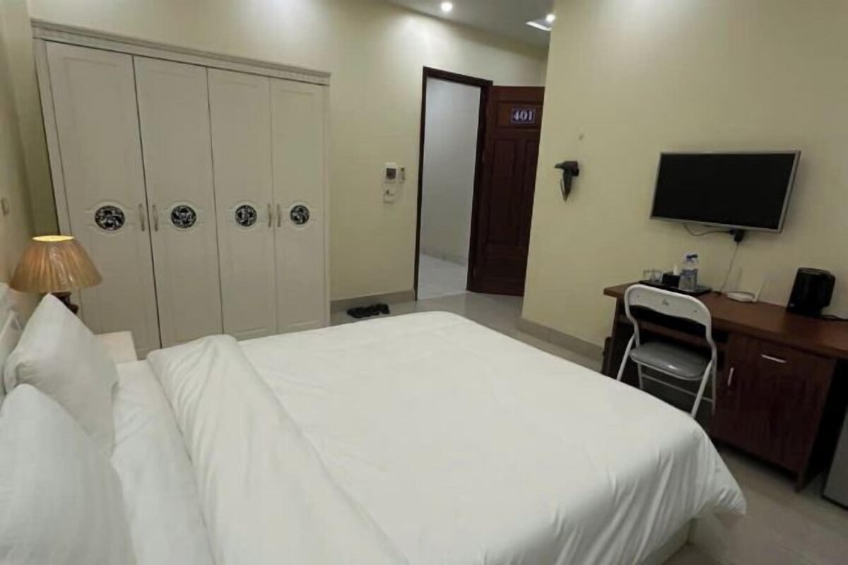 Quarto de hotel com cama, escrivaninha, cadeira, televisão e guarda-roupa.