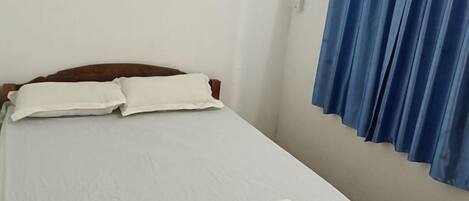 Egy egyszemélyes ágy fehér ágyneművel és párnákkal, egy fafejtámlával, és egy kék függönnyel az ablakon.