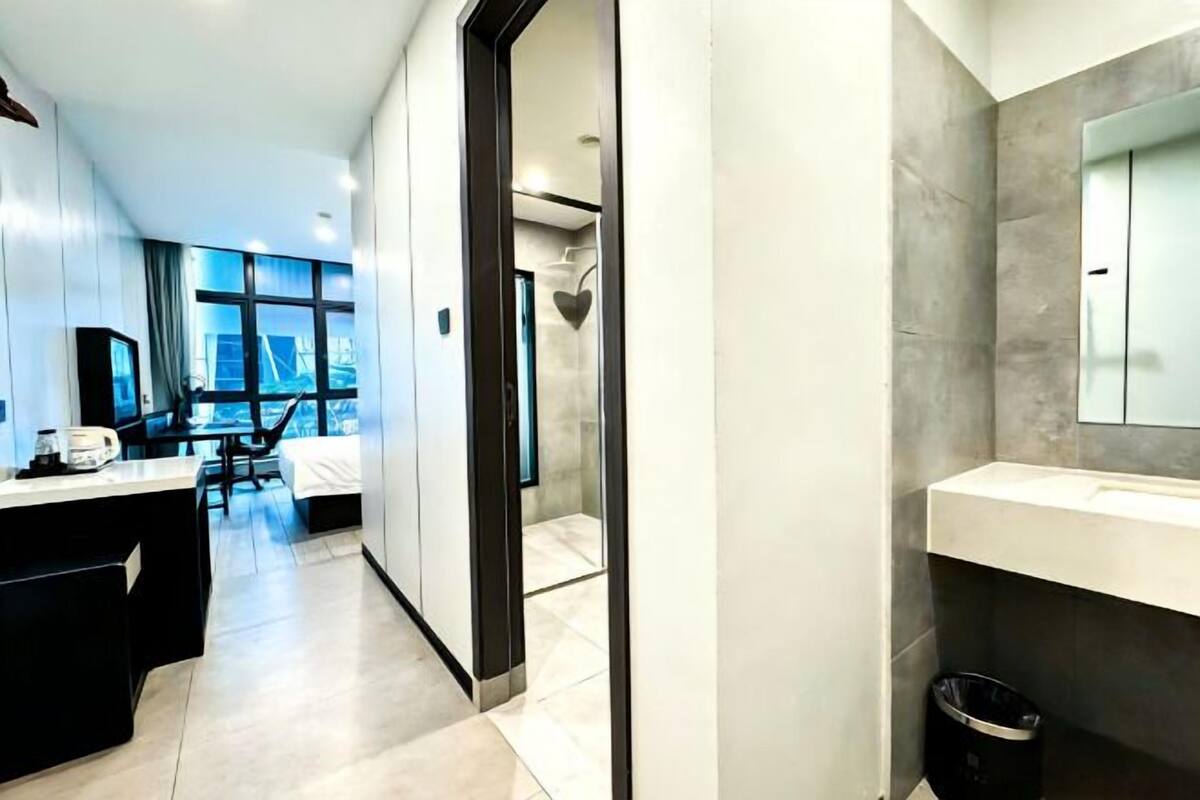 A modern hotel room with a large window, a desk, a chair, a bed, and a bathroom with a mirror and sink.