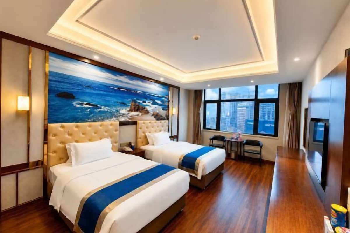 A hotel room with two beds, a large window, a city view, and a painting of the ocean.