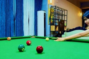A pool table with balls, a person leaning over it, and a room with curtains and a shelf with items.