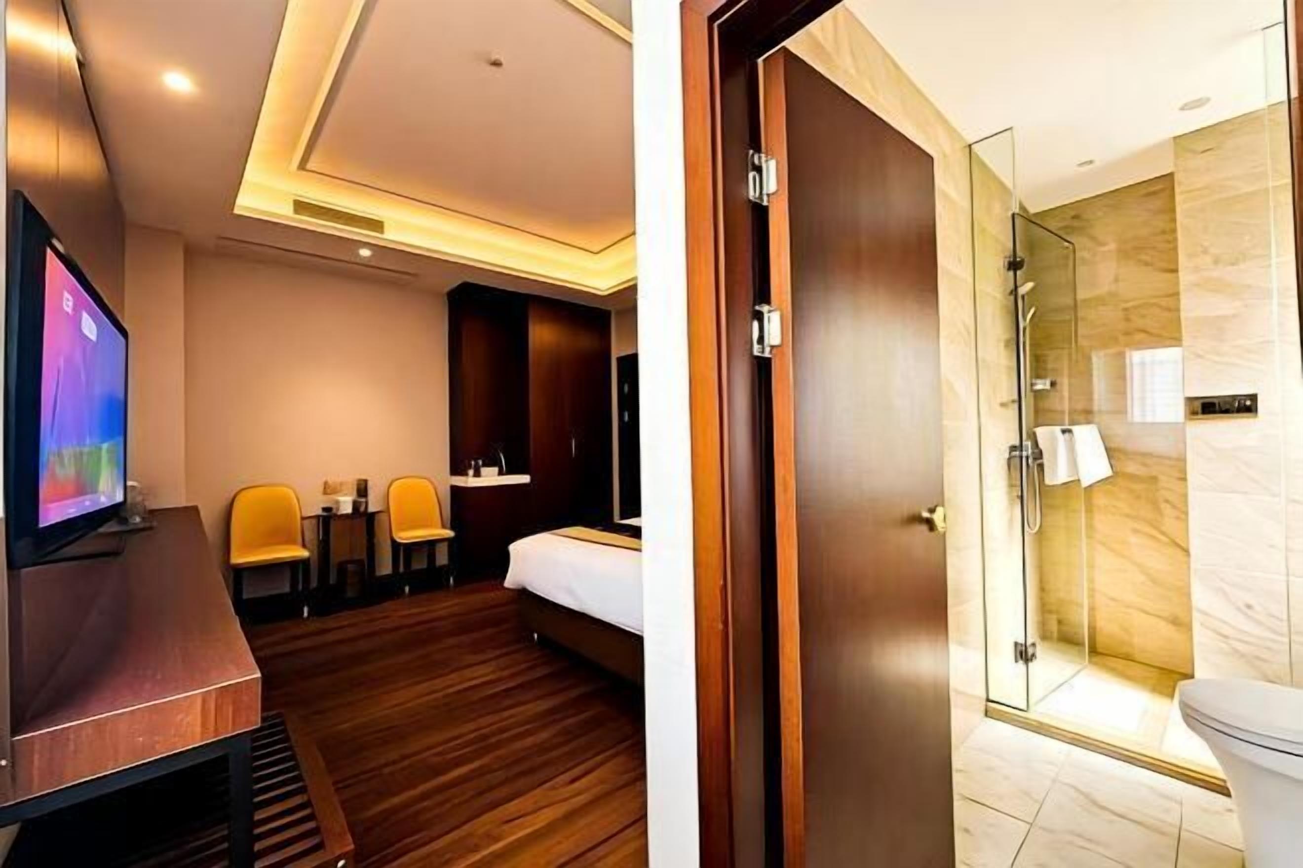 A hotel room with a TV, wooden flooring, a bed, a desk, and a shower area.