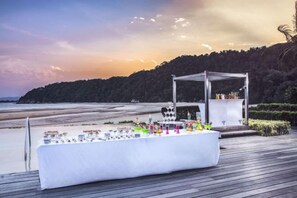 Montaje para un evento en la playa con una larga mesa cubierta de botellas y copas, una estructura de toldo blanco y una terraza de madera.