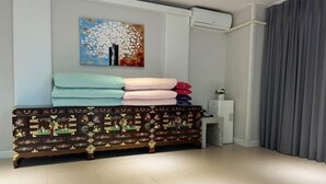 A room with a decorative wooden dresser, a wall-mounted air conditioning unit, a small white table with a vase, and a gray curtain.