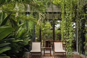 A wooden deck with two white cushioned chairs and a table, surrounded by lush green plants and a hanging garden structure. - Private Nongnia Beach Villas (Hoi An)